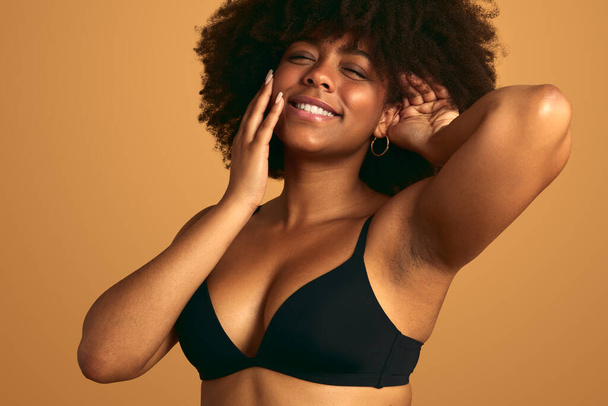 Portrait of cheerful young African American female model in black underwear with closed eyes, smiling and touching face and Afro hairstyle against beige background in studio - Foto, Bild