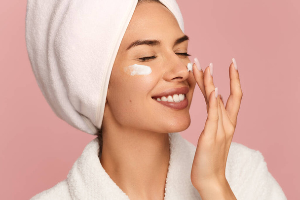 Positive young female model with perfect skin and towel on head smiling with closed eyes, while applying moisturizing cream on cheeks and nose after bath against pink background - Photo, image