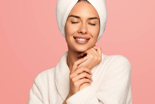 Smiling young female in white bathrobe with towel on head and hand under chin standing against pink background after skincare routine - Photo, image