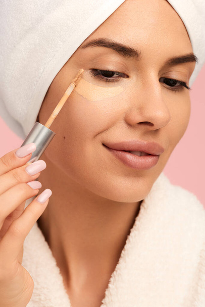 Young sensual female in white bathrobe with towel on head applying concealer with makeup brush under eye against pink background - Zdjęcie, obraz