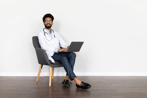 E-health, doctor appointment online, telemedicine concept. Happy smiling handsome young middle eastern man in workwear doc sitting in armchair, using laptop, isolated on white background, copy space - 写真・画像