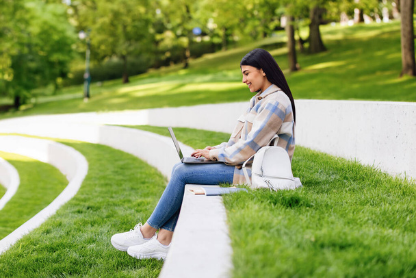 Distance learning concept. Happy hispanic student lady having remote lecture or browsing internet on laptop, sitting outdoors in park. E-learning, people and gadgets - 写真・画像