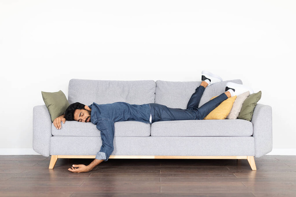 Exhausted tired young eastern man wearing casual outfit sleeping on couch at home over white blank wall. Drunk indian guy lying on sofa, experiencing hangover, copy space - Fotoğraf, Görsel