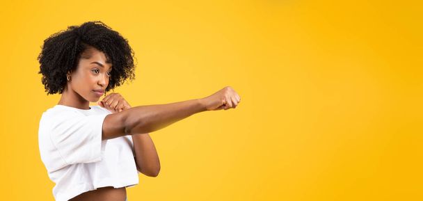 Σοβαρή αυτοπεποίθηση χιλιετή αφροαμερικάνικη σγουρή γυναίκα σε λευκό t-shirt καταπολέμηση, διάτρηση, απομονώνονται σε κίτρινο φόντο στούντιο, πανόραμα. Αυτοάμυνα, μάχη, δύναμη, δύναμη, διαφήμιση και προσφορά - Φωτογραφία, εικόνα