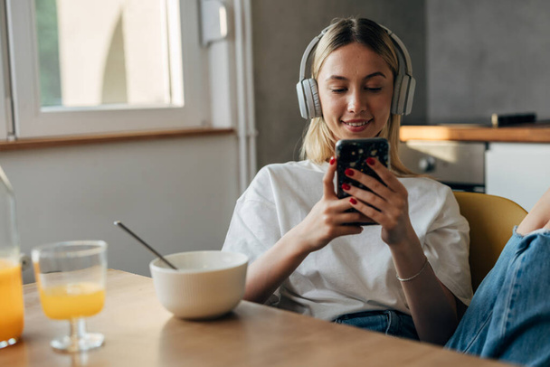Front view of a beautiful blond woman sitting in the kitchen and using her mobile phone in the morning. - Foto, afbeelding