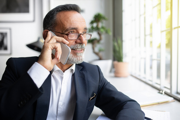 Confident european senior businessman in suit, glasses calls by phone at table, has meeting remotely in office interior. Work, business, create startup project and communication, close up - Photo, Image