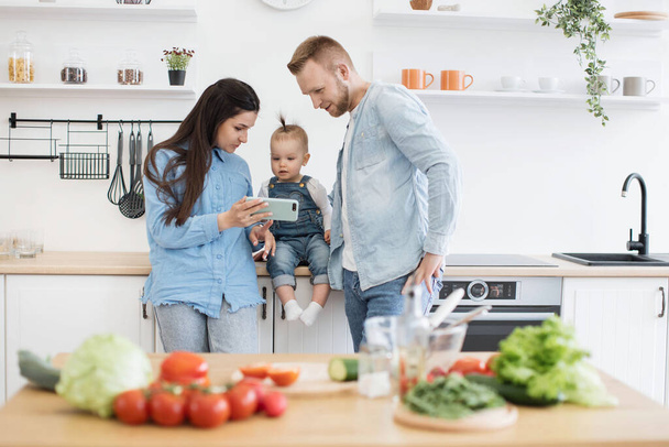 Happy young family of three in denim outfits looking at mobile screen behind kitchen table at home. Parents and kid searching for online recipe on phone with fresh ingredients for salad in foreground. - Foto, imagen