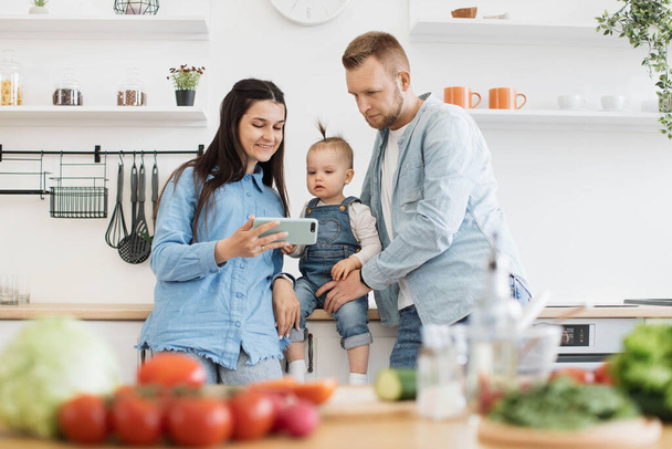 Attractive woman holding smartphone while mindful father looking at dining table with fresh products in modern kitchen. Loving spouses and little daughter watching cooking video online via gadget. - Fotó, kép