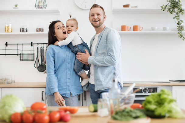 Portrait of joyful young parents in denim clothes cuddling little daughter while staying in kitchen of spacious apartment. Loving adult people and kid lifting mood while cooking together at home. - Foto, Imagem