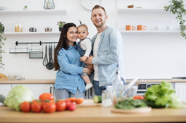 Portrait of joyful young parents in denim clothes cuddling little daughter while staying in kitchen of spacious apartment. Loving adult people and kid lifting mood while cooking together at home. - Fotografie, Obrázek