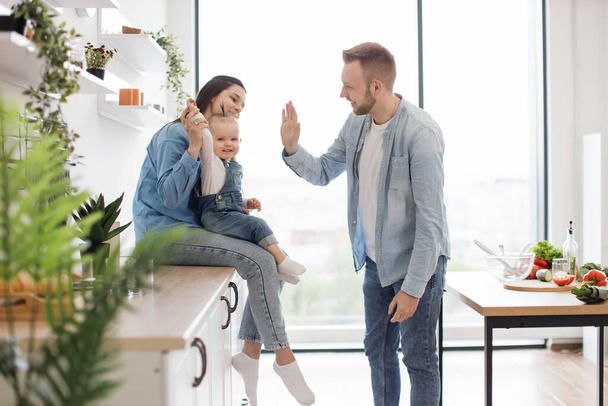 Beautiful brunette woman sitting on countertop with baby on laps while handsome man standing in middle of room. Funny father playing clapping game with daughter and mother in kitchen interior. - Photo, Image