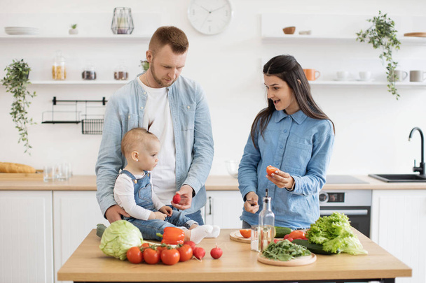 Charming brunette lady cutting red vegetable while mindful dad showing radish to little daughter on dining table. Adoring three-person family preparing healthy breakfast together in modern kitchen. - Фото, изображение