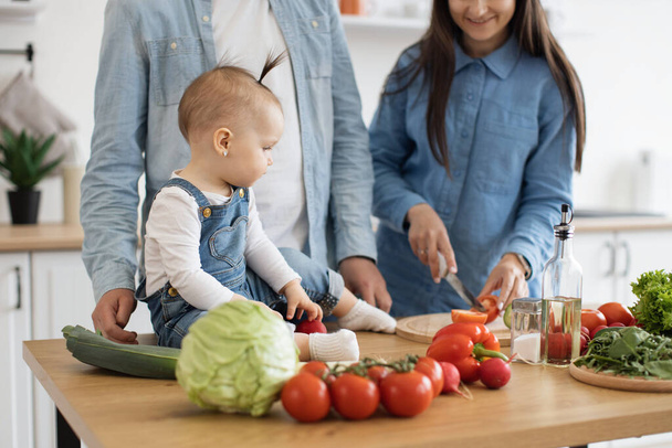 Cropped view of brunette female holding cucumber while sweet baby girl reaching out hand for green vegetable. Loving young mother teaching daughter eating healthy meal prepared together at home. - 写真・画像