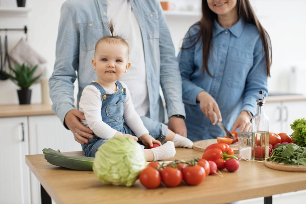 Close up view of adorable infant sitting on dining table among fresh crunchy products ready for garden salad. Cute baby daughter participating in family dinner preparation on sunny day at home. - Photo, image