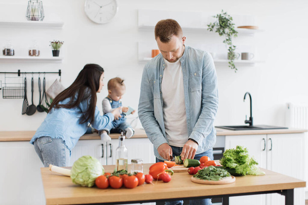 Attractive caucasian man slicing raw cucumber while brunette woman and baby girl using cell phone on kitchen worktop. Cheerful couple creating family recipes while preparing healthy dishes at home. - Foto, imagen