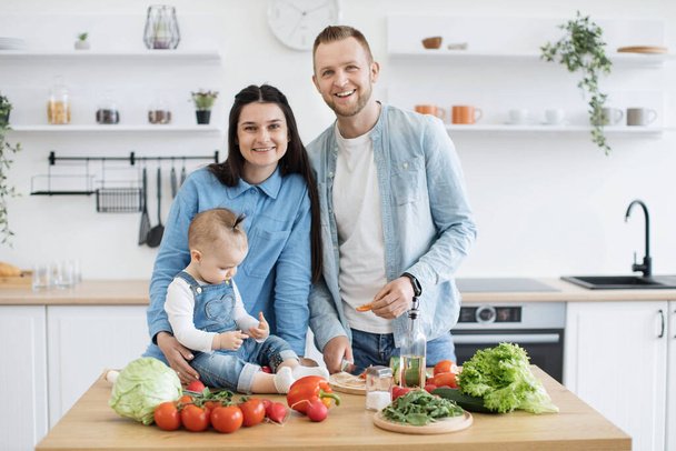 Portrait of happy three-person family in denim wear standing at kitchen table with assorted vegetables and greens. Stylish spouses achieving balanced diet by using organic products for home cooking. - 写真・画像