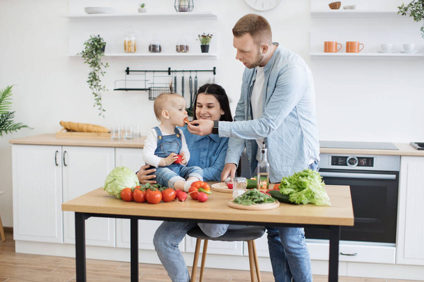 Delighted family man feeding little daughter while smiling mother sitting on kitchen chair and cheering baby girl on. Young spouses adding new nutrition to childs diet by offering sweet tomato. - Foto, immagini
