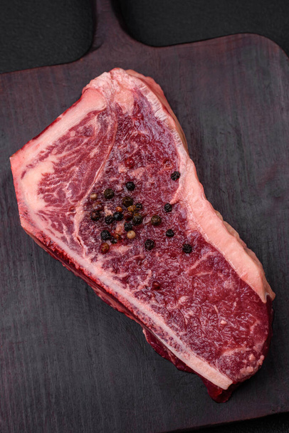 Čerstvé šťavnaté syrové hovězí steak z New Yorku se solí, kořením a bylinkami na tmavém betonovém pozadí - Fotografie, Obrázek