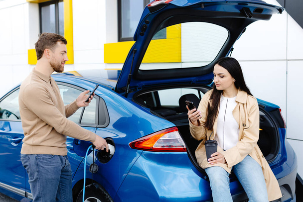 Young couple man and woman traveling by electric car having stop at charging station. Boyfriend plugging in cable to charge. Man talking with girlfriend, holding cup drinking hot coffee smiling. - Photo, Image