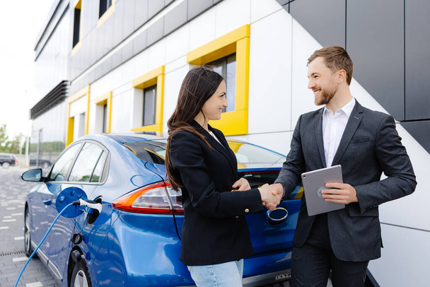 Smiling woman standing at the car and pointing at car seller tablet. She is picked right car for her she saw online. A woman concludes an agreement to buy a car and shakes the manager's hand. - Foto, Bild