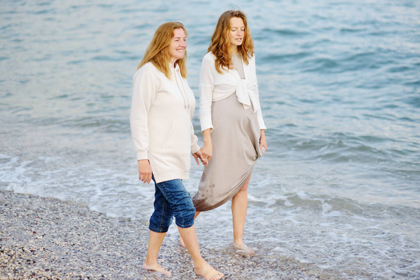 Redhead senior mother and her adult pregnant daughter are walking together on the sea shore. Happy loving mom and her grown up child talking and holding hands. - Foto, Bild