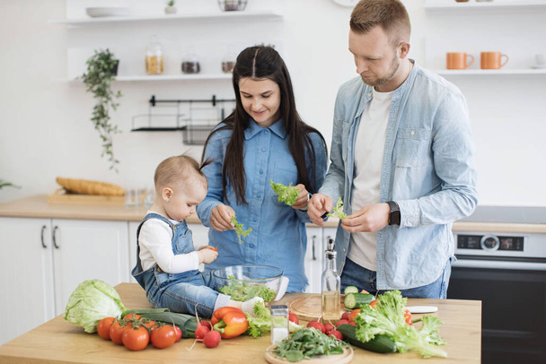 Cheerful spouses in cozy outfits putting leaves of fresh greens into bowl while small kid thinking about adding tomato. Young parents and daughter looking forward to healthy nutritious breakfast. - Foto, imagen