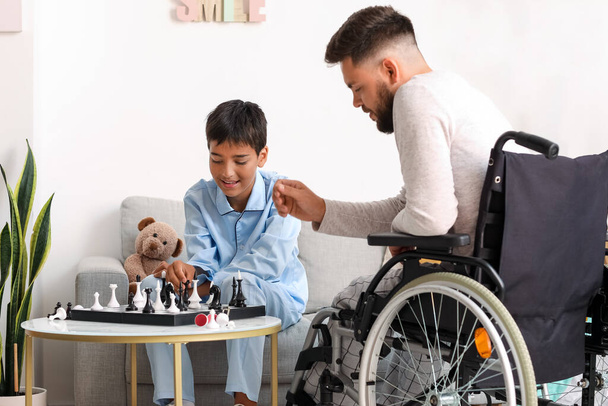 Маленький мальчик и его отец в инвалидной коляске играют в шахматы дома - Фото, изображение