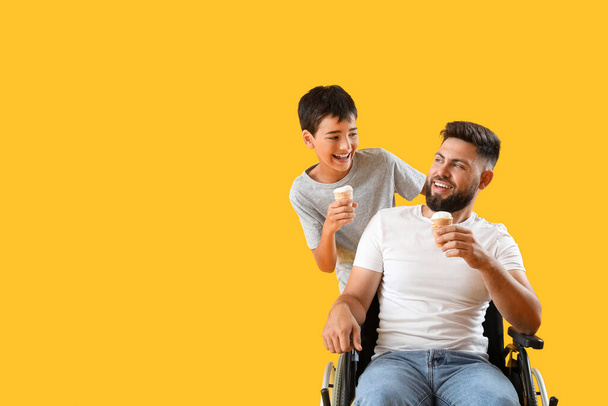 Μικρό αγόρι και ο πατέρας του σε αναπηρική καρέκλα με παγωτά σε κίτρινο φόντο - Φωτογραφία, εικόνα