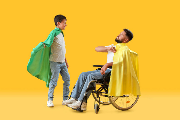 Malý chlapec a jeho otec na invalidním vozíku na sobě superhrdina pláště na žlutém pozadí - Fotografie, Obrázek