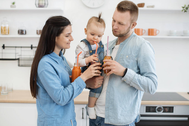 Young married couple with kid spending happy time together in cozy kitchen at home. Playful parents holding bottles with multivitamin juice and looking at baby with funny face expressions. - Фото, зображення
