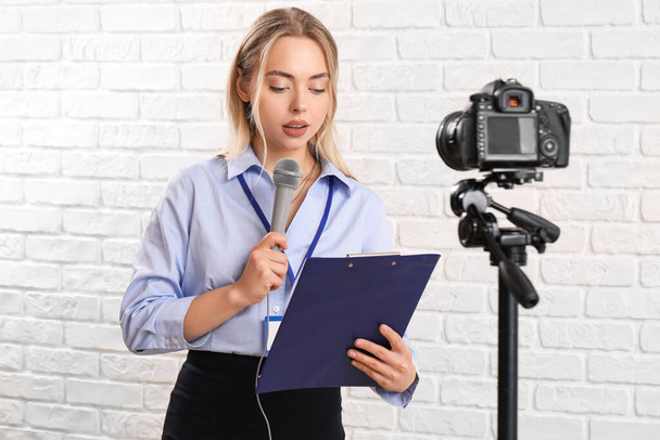Female journalist with microphone and clipboard recording video on white brick background - Photo, Image