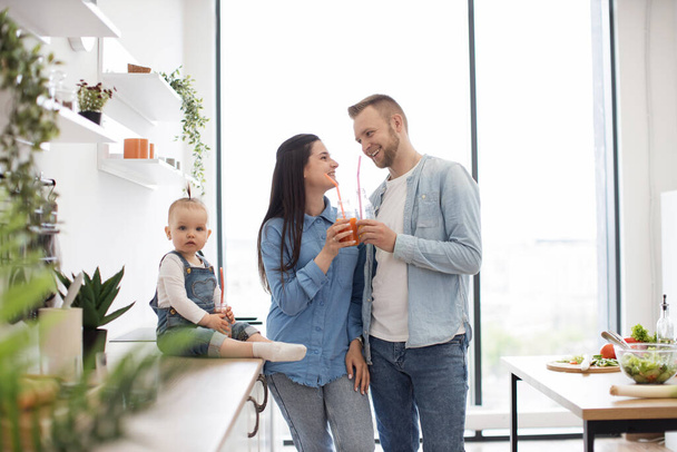 Affectionate husband and wife holding fruit juice in hands and looking at each other with smile indoors. Curious little toddler sitting on working surface next to parents and posing on camera. - Фото, изображение