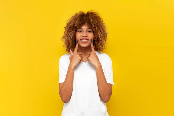 giovane ragazza riccia americana con bretelle punta il dito tra parentesi su sfondo giallo isolato, donna africana in t-shirt bianca mostra sorriso dentato e buon umore - Foto, immagini