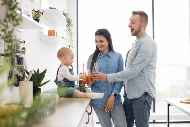 Adorable family dressed in denim attire toasting fresh drinks while resting in white spacious kitchen. Attentive parents taking care of little daughter while staying at home. Concept of love. - Foto, immagini