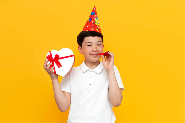 asian little boy of ten years old in party hat with gift celebrates birthday on yellow isolated background, korean child with heart-shaped gift congratulates - Photo, Image
