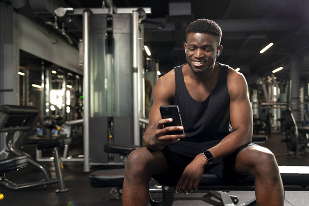 giovane atletico uomo africano americano si siede in palestra scura e utilizza smartphone, i tipi ragazzo sportivo online al telefono nel fitness club e sorride - Foto, immagini