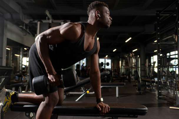 młody atletyczny amerykański mężczyzna trenuje w ciemnej siłowni, atletyczny facet podnosi ciężkie hantle w klubie fitness - Zdjęcie, obraz