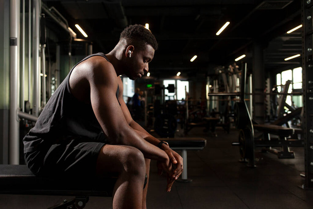 young athletic african american man sits in dark gym and thinks, pensive athletic guy rests and looks forward in fitness club, portrait of athlete in training - Foto, Bild