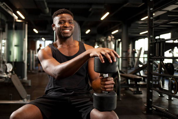 joven atlético afroamericano hombre está entrenando en el gimnasio oscuro y sonriendo, chico deportivo está sentado y sosteniendo pesadas mancuernas en el club de fitness, retrato de atleta en el entrenamiento - Foto, imagen