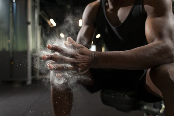sportovní afroameričan muž vlaky v tmavé tělocvičně a tře si ruce s magnézií, mladý chlap dělá tleskat s rukama v posilovně, detailní záběr prachu a mouky s rukama atleta - Fotografie, Obrázek