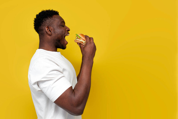 Afrykański Amerykanin w białej koszulce trzyma wielkiego burgera z otwartymi ustami, facet je fast food z boku na żółtym, odizolowanym tle, przestrzeń na ksero - Zdjęcie, obraz