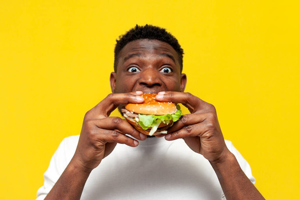 shocked african american man in white t-shirt holding big burger and surprised, amazed guy eating fast food on yellow isolated background, emotions from delicious food - Photo, Image