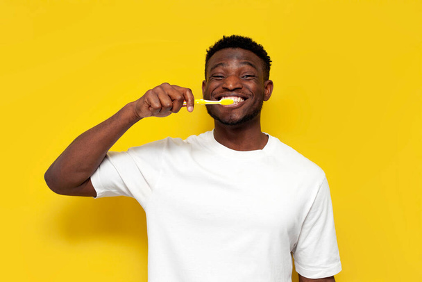 アフリカ系アメリカ人の男が歯ブラシで歯をブラッシング黄色の隔離された背景で男は歯と笑顔の世話をします - 写真・画像
