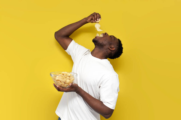 happy african american man holding plate of chips and eating snacks from potatoes on yellow isolated background, guy in white t-shirt eats fast food - Photo, Image