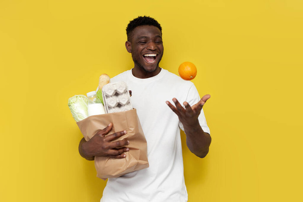 gelukkig Afrikaans amerikaans man shopper holding eco tas met boodschappen en knikken oranje tegen geel geïsoleerde achtergrond, guy klant verheugen en glimlachen - Foto, afbeelding
