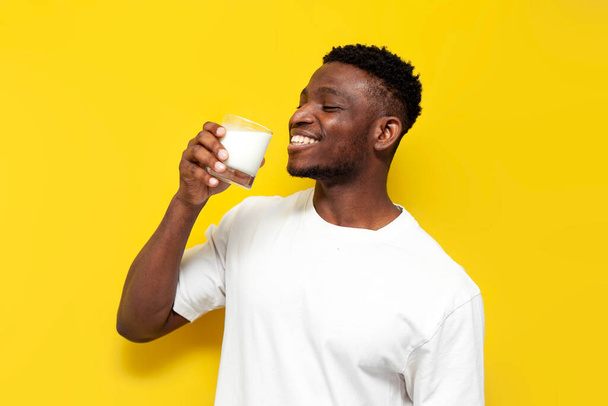 Beyaz tişörtlü Afro-Amerikan adam sarı izole edilmiş arka planda bardakta süt içiyor, elinde taze beyaz süt ürünü bardakları tutuyor ve gülümsüyor. - Fotoğraf, Görsel