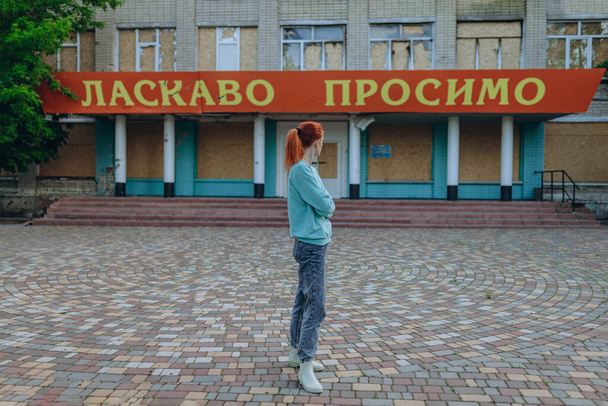 Bayan bomba patlamasından sonra köydeki hasarlı ve tahrip edilmiş okulun önünde duruyor. Girişte "Hoş geldiniz" yazıyor. Ukrayna 'ya karşı savaş. Rus bombası okulu vurdu. Terör saldırısı. Kötülük - Fotoğraf, Görsel