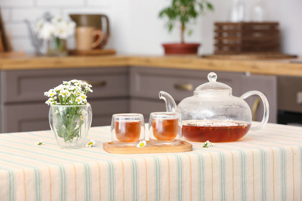 Drewniana taca z czajnikiem, kubki naturalnej herbaty rumianku i kwiaty na stole w kuchni - Zdjęcie, obraz