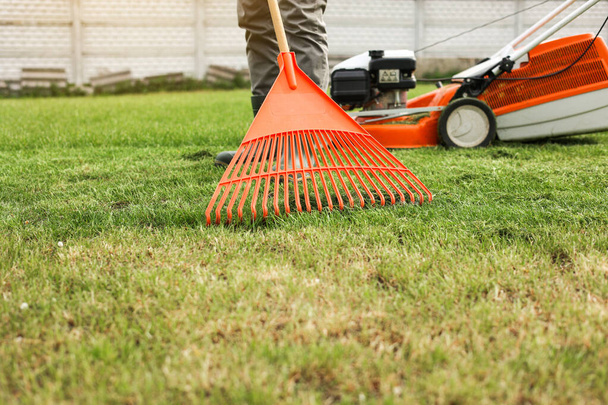 Male gardener collects cut grass with orange plastic rake after a mower, works in the backyard of the house. Man takes care of lawn. Concept of housework, gardening and country life, garden tools - Фото, изображение