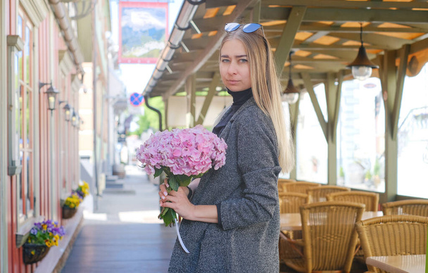 ブロンドの髪をした美しい白人の女の子、グレーのコートとピンクのアジサイの花束と背景に街の景色を望むサングラス付きの青いジーンズ. - 写真・画像